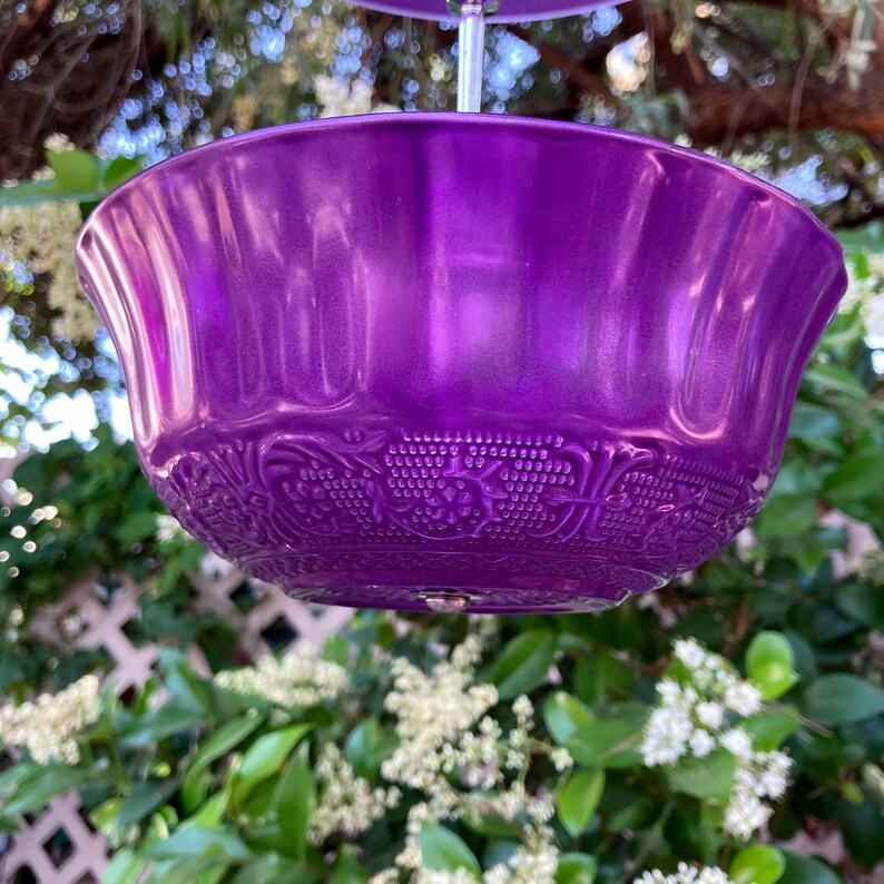 May include: A vibrant purple bowl with a glossy finish. The bowl features a decorative embossed pattern around the lower edge. The bowl is suspended in front of a background of green foliage and a white trellis.
