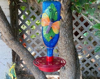 May include: A blue hummingbird feeder with a butterfly design. The feeder has a red base and a decorative butterfly with orange wings and a purple body. The feeder is hanging from a tree branch.