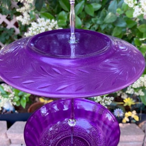 May include: A two-tiered serving tray with a metallic purple finish. The top tier features a leaf pattern, while the bottom tier has a detailed floral design. The tray is supported by a silver metal stand, perfect for displaying desserts or appetizers.