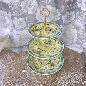 May include: A three-tiered serving stand with yellow plates featuring floral and Chinese character designs. The plates have a blue and green border and are connected by a gold-colored handle. The stand is likely used for displaying desserts or appetizers.