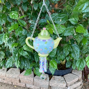 May include: A whimsical, hanging teapot wind chime. The ceramic teapot is light blue with purple and pink floral accents, a green lid, and a yellow handle and spout. It hangs from a silver chain and features beaded strands and a clear crystal ball.