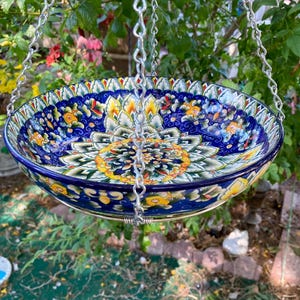 May include: A hanging ceramic bird bath with a detailed floral design. The bowl is primarily blue with yellow, white, and green accents. It is suspended by a silver chain, creating a decorative garden element.