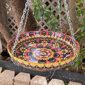 May include: A round, shallow ceramic bird feeder or hanging bowl featuring a vibrant, multi-colored mandala pattern with yellow, red, green, and black floral motifs. The bowl is suspended by three silver-toned metal chains attached to the rim.