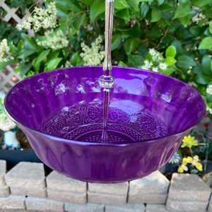 May include: A vibrant purple glass serving bowl with a decorative embossed pattern. The bowl is suspended by a silver metal handle with a decorative jewel accent. The bowl is designed for serving food or displaying decorative items.