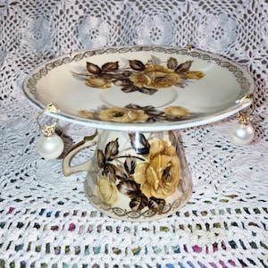 May include: A white porcelain teacup and saucer with a floral design, used as a decorative earring holder. The cup and saucer are decorated with brown and yellow roses.
