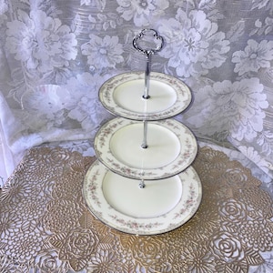 May include: A three-tiered serving stand with a silver metal frame and three white ceramic plates with a delicate floral pattern. The plates have a gold trim and a subtle pink and green floral design.