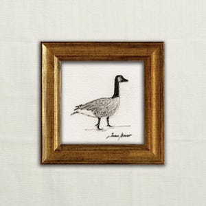 May include: A framed pencil sketch of a Canada goose. The artwork depicts a detailed drawing of the bird with a black head and neck, grey body, and black legs. The artwork is in a gold-coloured frame.