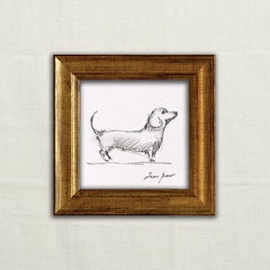 May include: A framed pencil sketch of a dachshund dog. The artwork is in a gold-colored, square frame. The dog is drawn in profile, with its head and tail raised. The artist's signature is visible at the bottom.