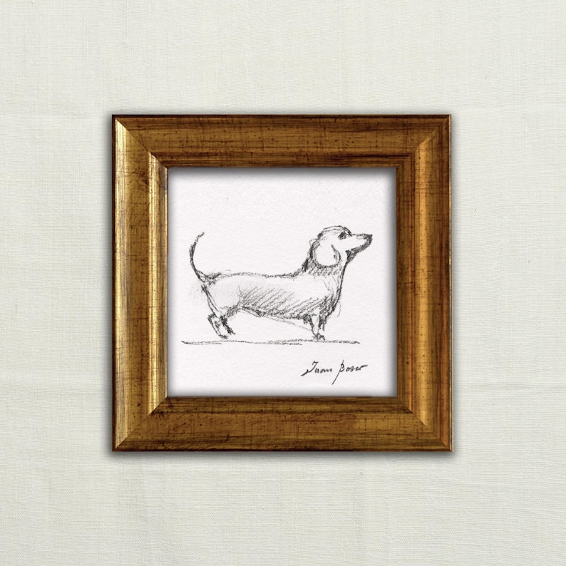 May include: A framed pencil sketch of a dachshund dog. The artwork is in a gold-colored, square frame. The dog is drawn in profile, with its head and tail raised. The artist's signature is visible at the bottom.