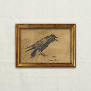 Op de afbeelding: Een ingelijste houtskooltekening op beige papier met een zijaanzicht van een kraai met een open snavel. De vogel is getekend met donkere, expressieve lijnen. Het kunstwerk zit in een eenvoudige, goudkleurige metalen lijst met een rechthoekig profiel.
