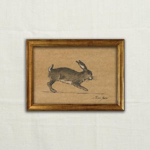 May include: A framed charcoal drawing on tan paper depicting a running hare in profile. The artwork is enclosed in a rectangular gold-toned frame with a textured finish. A handwritten signature is visible in the bottom right corner of the drawing.