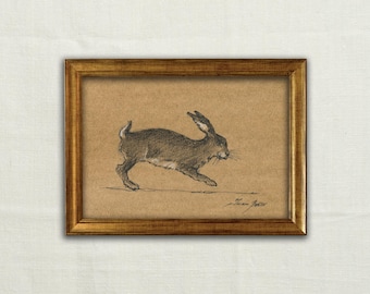 May include: A framed charcoal drawing on tan paper depicting a running hare in profile. The artwork is enclosed in a rectangular gold-toned frame with a textured finish. A handwritten signature is visible in the bottom right corner of the drawing.