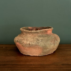 May include: A small, round, terracotta pot with a rough, textured surface. The pot is a reddish-brown colour with patches of green and grey.