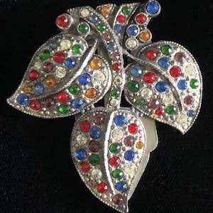 Puede incluir: Un broche plateado con un diseño de tres hojas, cada una cubierta de piedras de cristal de colores. Las piedras de cristal son en tonos de rojo, azul, verde y transparente.