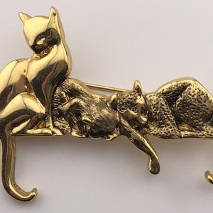 Könnte beinhalten: Goldfarbene Brosche mit drei stilisierten Katzen. Eine Katze sitzt aufrecht, während die anderen beiden liegen. Die Brosche hat einen Nadelverschluss. Die Katzen haben detaillierte Fellstrukturen und gebogene Schwänze.