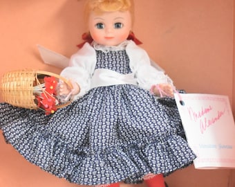 May include: A Madame Alexander doll with blonde hair, wearing a white long-sleeved top, a blue and white floral print dress with a ruffled hem, red tights, and black shoes. The doll holds a small woven basket and includes a white tag with text.