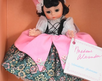May include: A Madame Alexander doll with dark hair, wearing a pink bonnet with white lace, a white blouse, a black bodice, and a skirt featuring a floral pattern in green, pink, and yellow. A pink overskirt and a white tag with pink text are included.