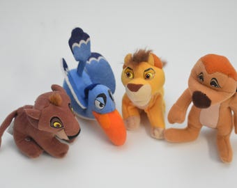 May include: A collection of plush toys featuring characters from the animated film, The Lion King. The toys include a brown lioness, a blue bird with an orange beak, a yellow lion, and a tan meerkat.