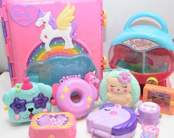 May include: A collection of Polly Pocket plastic toy cases in various shapes and colors, including a large pink rectangular case with a unicorn and rainbow, a blue and pink gumball machine case, and several smaller animal and food-themed compacts.