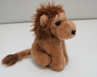 May include: A small, light brown plush lion toy with a soft, fuzzy texture. The stuffed animal features a darker brown, shaggy mane around its head, two small rounded ears, black plastic eyes, a black stitched nose, and a long tail with a tuft.