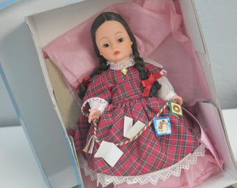 May include: A doll with dark hair in two braids, wearing a red and green plaid dress with white lace trim at the collar and hem. The doll is inside a box lined with pink tissue paper and holds a string of small decorative cards and white paper tags.
