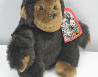 May include: A plush gorilla toy with dark brown fur, a lighter brown face, hands, and feet. It features a small pink ribbon on its head and a red Busch Gardens tag attached to its chest, displaying illustrations of various animals and the park name.