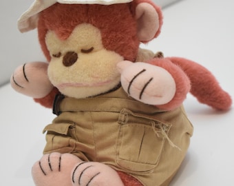 May include: A plush monkey toy with reddish-brown fur and a cream-colored face. The monkey wears a beige fabric safari hat and matching khaki cargo shorts with visible pockets. The toy has closed eyes, a brown nose, and stitched details on its paws.