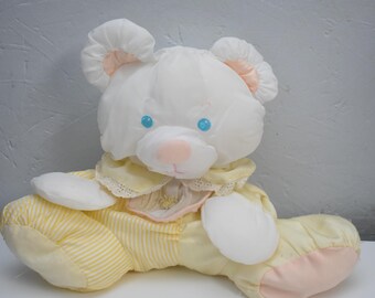 May include: A plush bear toy with white fur, bright blue eyes, and a soft pink nose. The bear wears a yellow ruffled collar with lace trim and yellow and white striped pants. The paws are white, and the feet have pink accents on the soles.