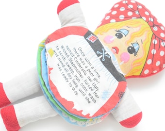May include: A fabric doll featuring a red and white polka dot head covering, yellow hair, and a printed story on its skirt. The text reads: Once upon a time, there was a poor girl named Cinderella. Her Ugly Wicked Stepmother forced Her to scrub, sweep, iron, cook, wash and mend all day long, until she was ready to drop.