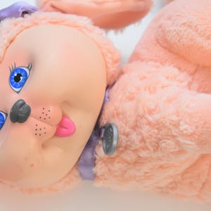 May include: A vintage pink plush dog toy with a plastic face. The dog has blue eyes, a gray nose, and a pink tongue sticking out. It has a purple bow and a silver button on its side.