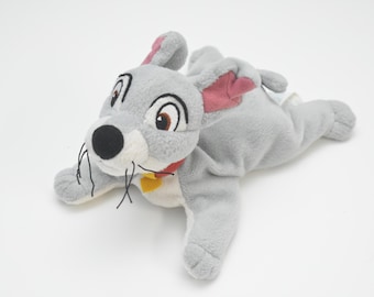 May include: A plush toy of a grey dog with large brown eyes, pink inner ears, and a black nose. The dog wears a red collar with a yellow heart-shaped tag and has black thread whiskers. The soft toy is in a lying position with its front paws extended.