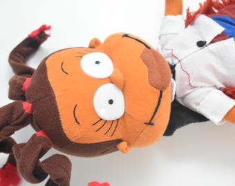 May include: A plush doll featuring a character with brown skin, large white eyes, and dark brown hair styled in multiple ponytails with red ties. The doll wears a white collared shirt with a black button, blue pants, and has red yarn accents.