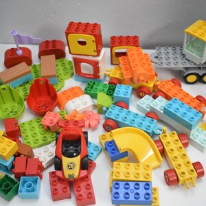 May include: A collection of colorful toy building blocks in various shapes and sizes. The blocks are primarily red, yellow, green, blue, and orange. There is a red race car with the number 28 on it, and a yellow slide. The blocks are scattered on a white surface.