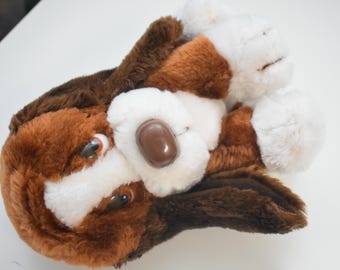 May include: A plush stuffed animal in the likeness of a basset hound dog. The toy features soft fur in shades of dark brown, light brown, and white. It has large, drooping ears, a smooth brown plastic nose, and brown eyes with white accents.