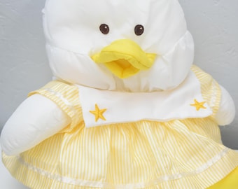 May include: A plush duck toy featuring a white head with a yellow beak, small black eyes, and a tuft of white yarn on top. The duck wears a white sailor-style collar with two yellow embroidered stars and a yellow and white striped dress.