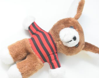 May include: A plush stuffed animal featuring brown fur with white accents on the ears, hands, and feet. The toy is wearing a shirt with horizontal red and black stripes. It has dark eyes and a stitched nose, with long ears extending from the head.