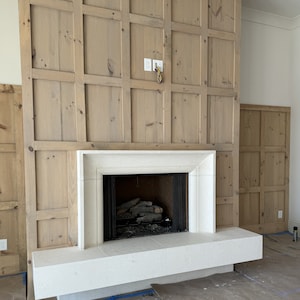May include: A white fireplace with a black metal grate and a wood-burning fire. The fireplace is surrounded by a light brown wood paneling.