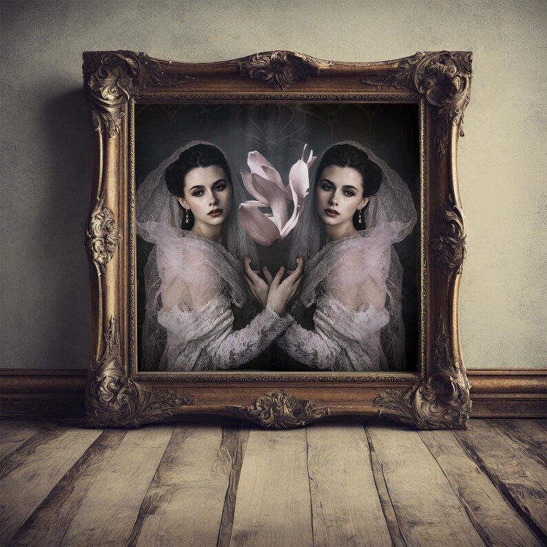 May include: A framed portrait featuring two women in vintage attire, holding a large pink flower. The ornate gold frame sits on a wooden floor, creating a classic and artistic aesthetic. The women are wearing white lace dresses and veils.