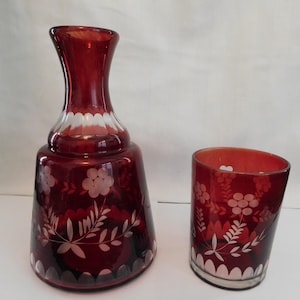 May include: A deep red glass carafe and a matching tumbler glass, both featuring etched white floral and leaf patterns along with a scalloped border design near the base and neck. The glass pieces have a smooth, translucent finish.