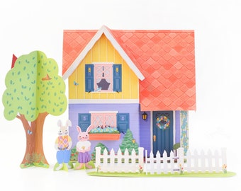 May include: A papercraft scene with a yellow and purple house with a red roof, a white picket fence, a green tree, and two bunny rabbits standing in front of the house.