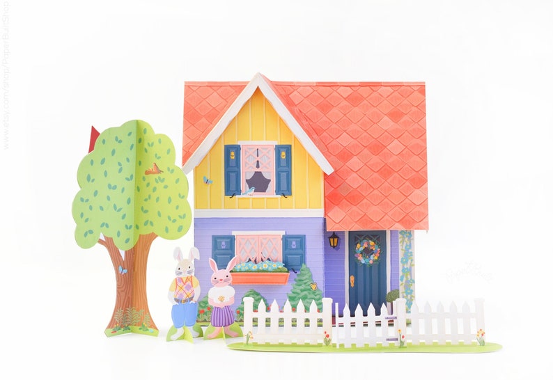 May include: A papercraft scene with a yellow and purple house with a red roof, a white picket fence, a green tree, and two bunny rabbits standing in front of the house.