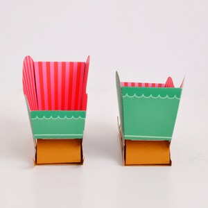 May include: Two paper boxes with different designs. The larger box is green with pink and white stripes on the top and a gold bottom. The smaller box is green with a scalloped edge and a gold bottom.