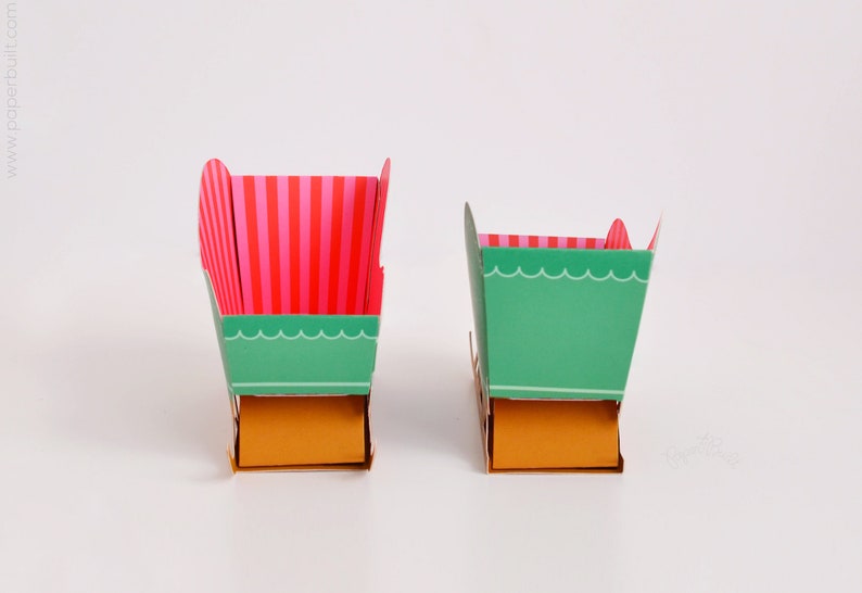 May include: Two paper boxes with different designs. The larger box is green with pink and white stripes on the top and a gold bottom. The smaller box is green with a scalloped edge and a gold bottom.
