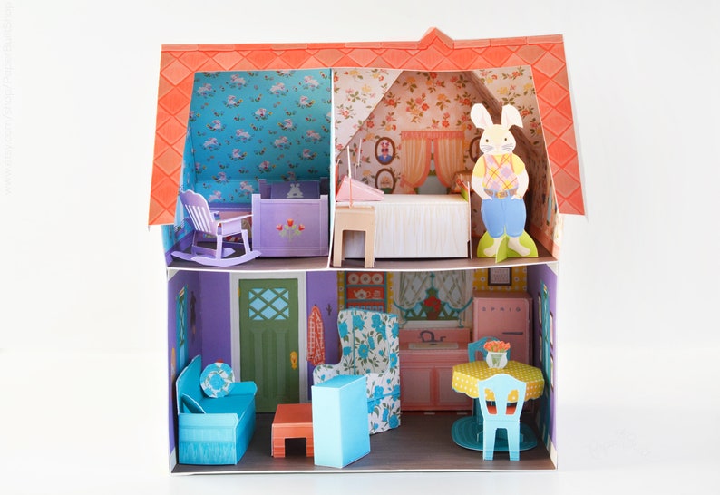 May include: A paper dollhouse with a purple exterior and a red roof. The dollhouse has two levels, each with a different room. The top level has a bedroom with a bed, a rocking chair, and a crib. The bottom level has a living room with a couch, a coffee table, and a chair. There is also a kitchen with a table and chairs.