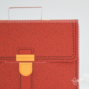 May include: Red leather-look briefcase with a handle and a clasp that says "PAPER BUILT".