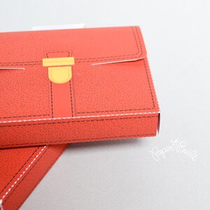 May include: A red cardboard box shaped like a briefcase with a yellow clasp and black stitching. The box is closed and the top is visible.