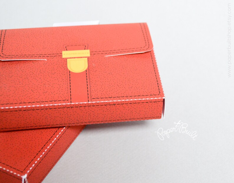 May include: A red cardboard box shaped like a briefcase with a yellow clasp and black stitching. The box is closed and the top is visible.