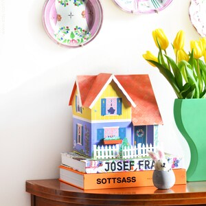 May include: A colorful paper dollhouse with a red roof sits on top of two books. The books are titled "Josef Frank" and "Sottsass". A white bunny figurine sits on the books. A vase with yellow tulips sits on a wooden table.
