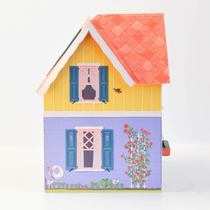 May include: A yellow and purple papercraft house with a red roof and green shutters. The house has a small garden with a red rose bush and a white picket fence.