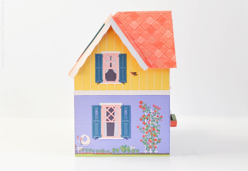 May include: A yellow and purple papercraft house with a red roof and green shutters. The house has a small garden with a red rose bush and a white picket fence.